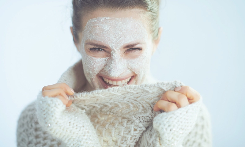happy modern woman in roll neck sweater and cardigan with white facial mask as part of winter skin care playing with clothes on winter light blue background.